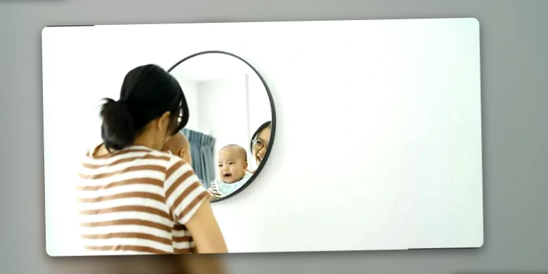 A mother holds her smiling baby in front of a round mirror, both looking at their reflection
